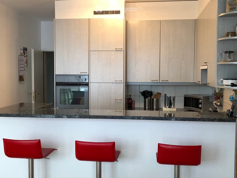 Modern kitchen with light wood cabinets, granite countertop, and three red bar stools. Appliances include an oven, microwave, and various kitchen utensils.