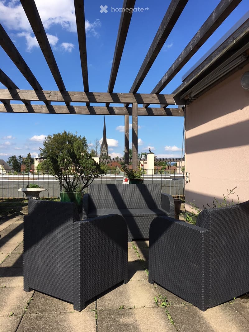 Wooden pergola-covered balcony with wicker sofa and chairs, overlooking a town with a church spire in the background.