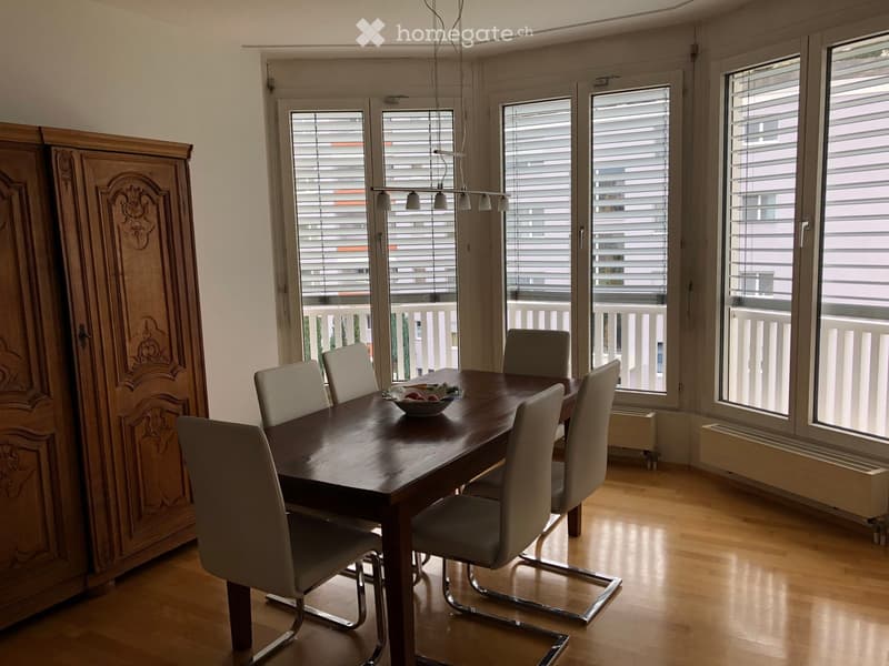 Bright dining room with wooden floor, large windows, and a balcony view. Features a wooden dining table with six chairs and a decorative wooden cabinet.