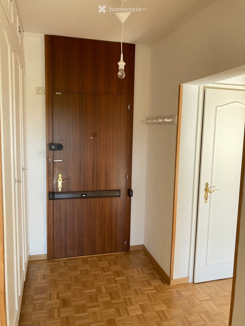 Empty hallway with wooden flooring, white walls, and two doors (one wooden, one white). A small shelf is mounted on the right wall, and a hanging light fixture is visible.