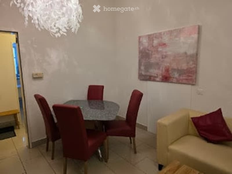 A small, modern dining and sitting area with a round glass dining table, red chairs, a yellow sofa, and a colorful abstract painting on the wall.