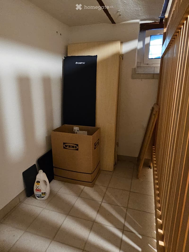 Empty hallway with tiled floor, wooden railing, and a small window letting in light. A cardboard box and cleaning supplies are placed against the wall.