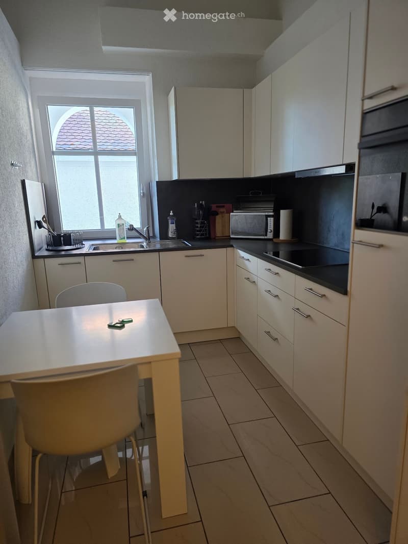 Modern kitchen with white cabinets, black countertops, a small dining table with a chair, and various appliances including a microwave and oven.