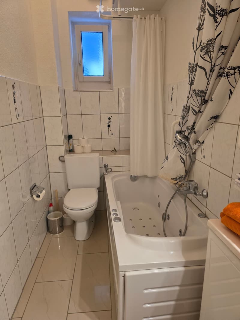 Bathroom with a bathtub, toilet, shower curtain, and a window above the bathtub. Toilet paper and a trash can are visible.