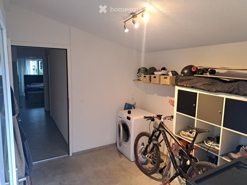 A small, well-lit room with a washing machine, a bicycle, shelves holding helmets and a skateboard, and a loft bed with storage compartments.