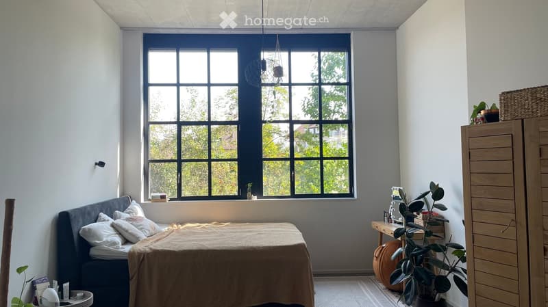 Modern bedroom with a large window, a bed with pillows and a blanket, a nightstand with a lamp, a wardrobe, a small table with a plant, and additional decor items.