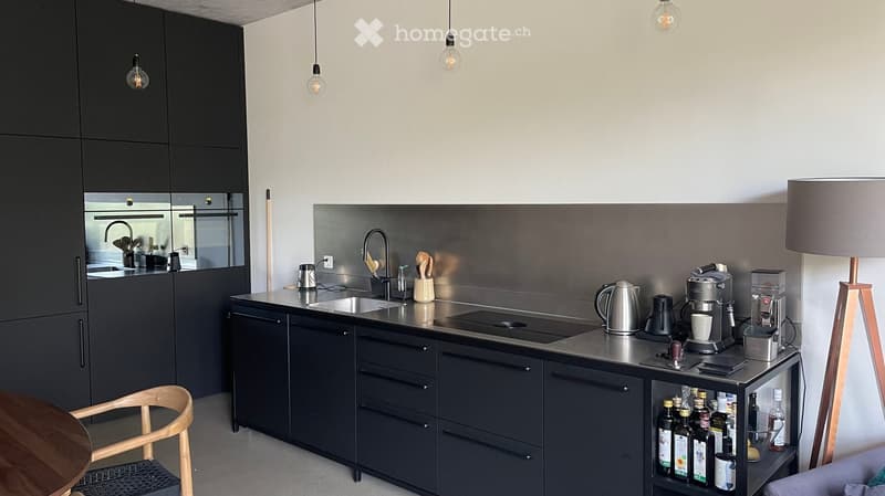 Modern kitchen with dark grey cabinets, stainless steel countertop, and various appliances including a coffee maker, kettle, and sink.
