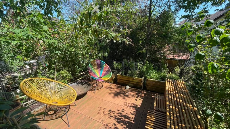 A wooden deck surrounded by lush greenery, featuring two colorful chairs and a small table, with wooden planters and a small staircase leading up.