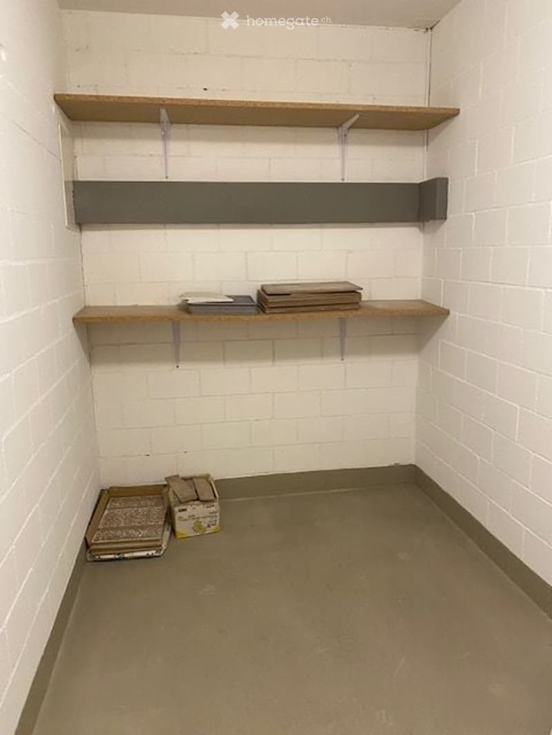 Empty storage room with wooden shelves, white brick walls, and concrete floor. Contains folded blankets and boxes on the floor.
