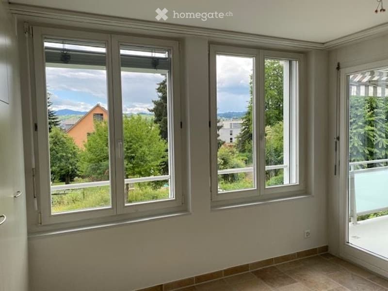Empty room with large windows offering a scenic view of trees and mountains, tiled floor, and a sliding glass door leading to a balcony.