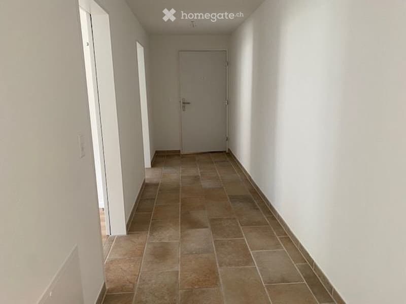 Empty hallway with tiled floor and white walls, featuring a closed door at the end and a light switch on the left wall.