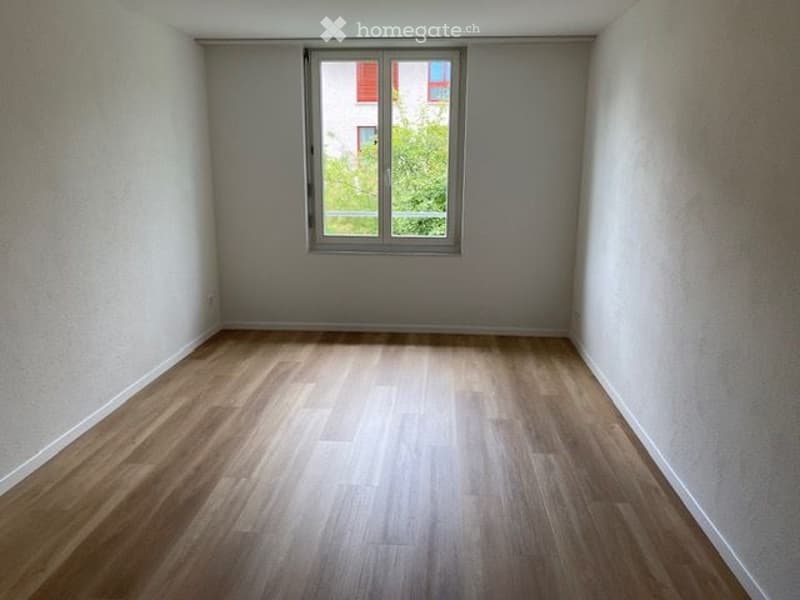 Empty room with light wooden flooring and a single window showing a view of greenery and a building outside.