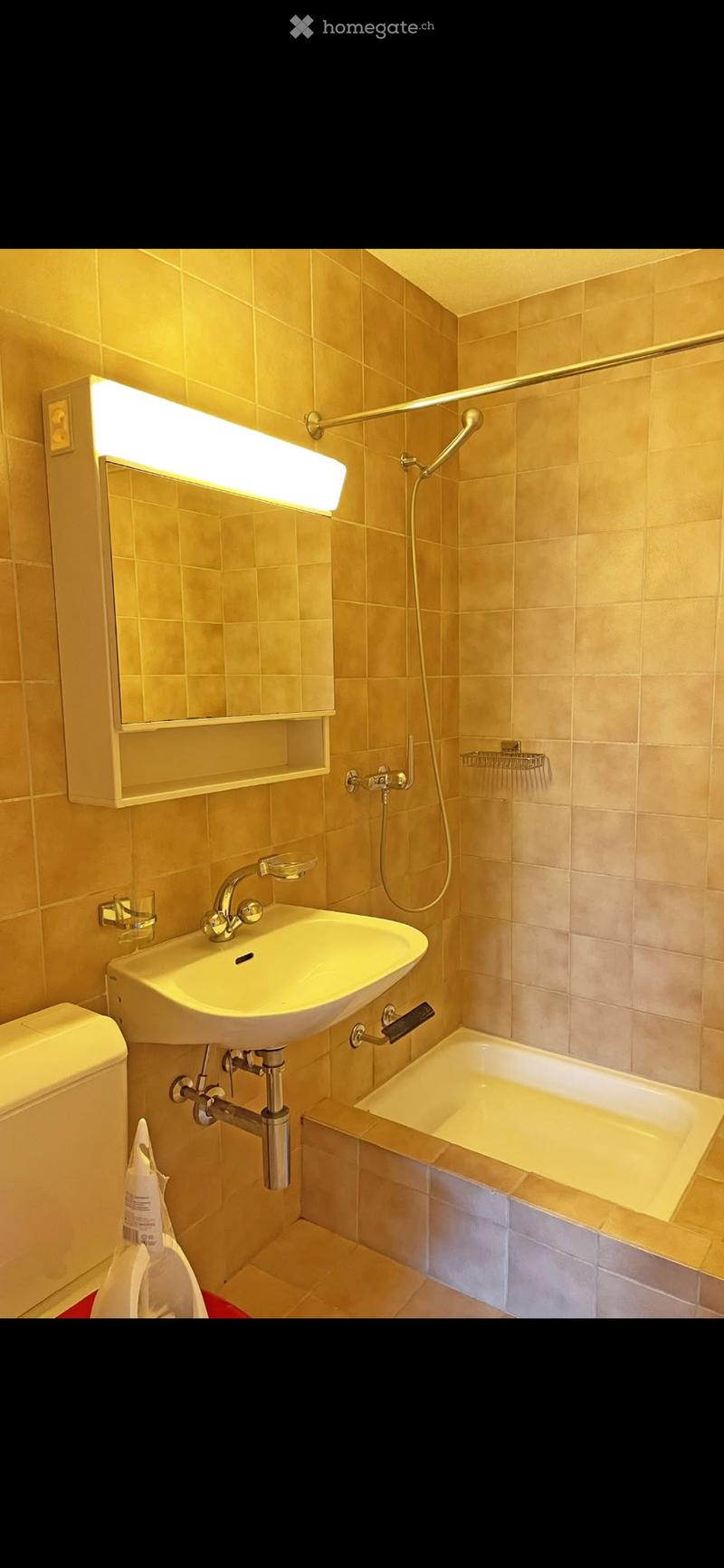 Bathroom with beige tiles, a white sink with a chrome faucet, a bathtub with a shower hose, a mirror with built-in lighting, and a toilet.