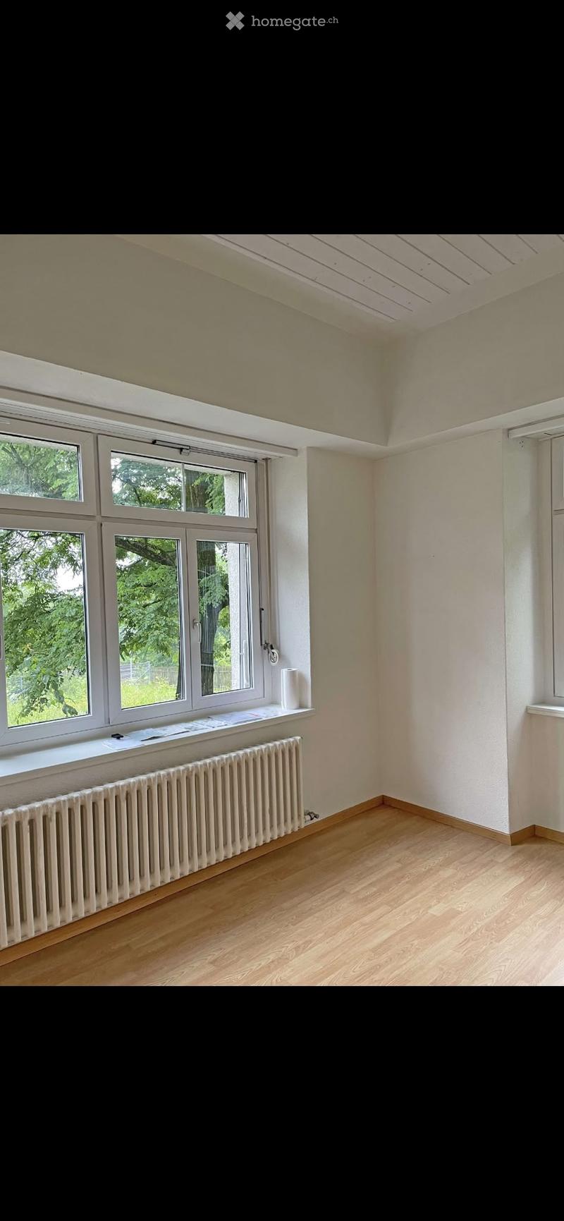 Empty room with wooden flooring, white walls, large windows, radiator, and a small shelf with a roll of paper towels and some papers.