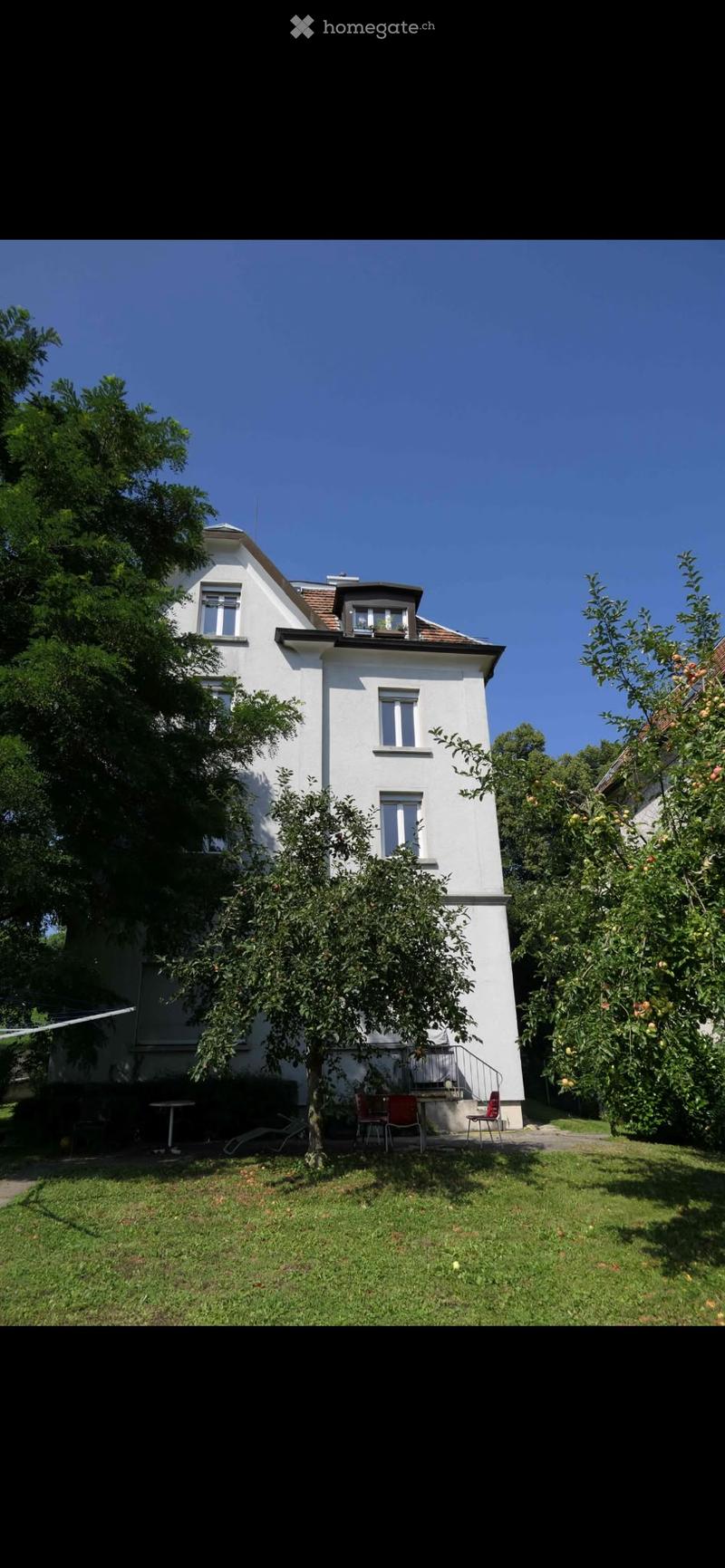 A two-story white house with a pitched roof, surrounded by a well-maintained garden with fruit trees and outdoor seating.