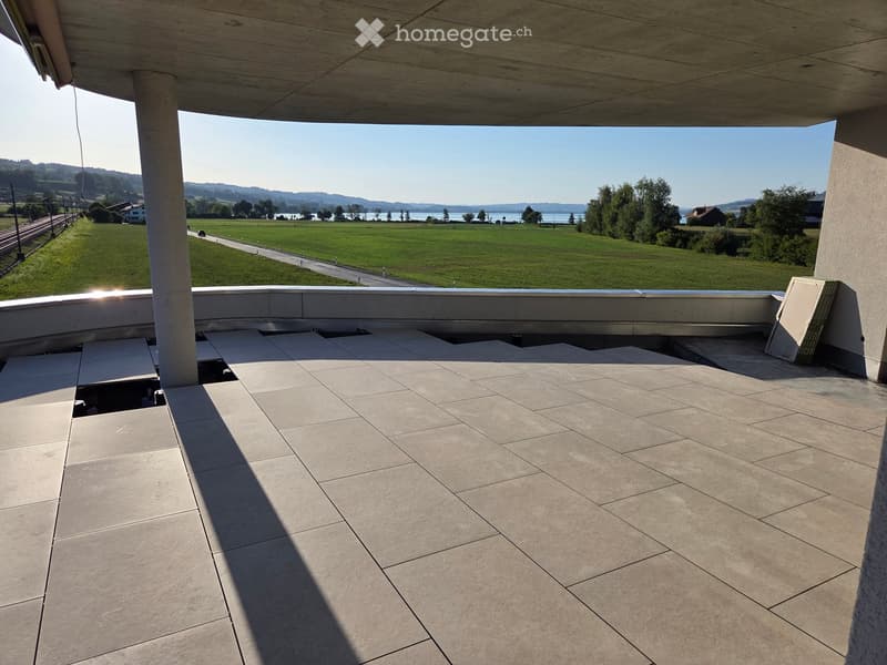 Modern balcony with tiled floor, concrete railing, and expansive view of a green field, road, and distant lake.