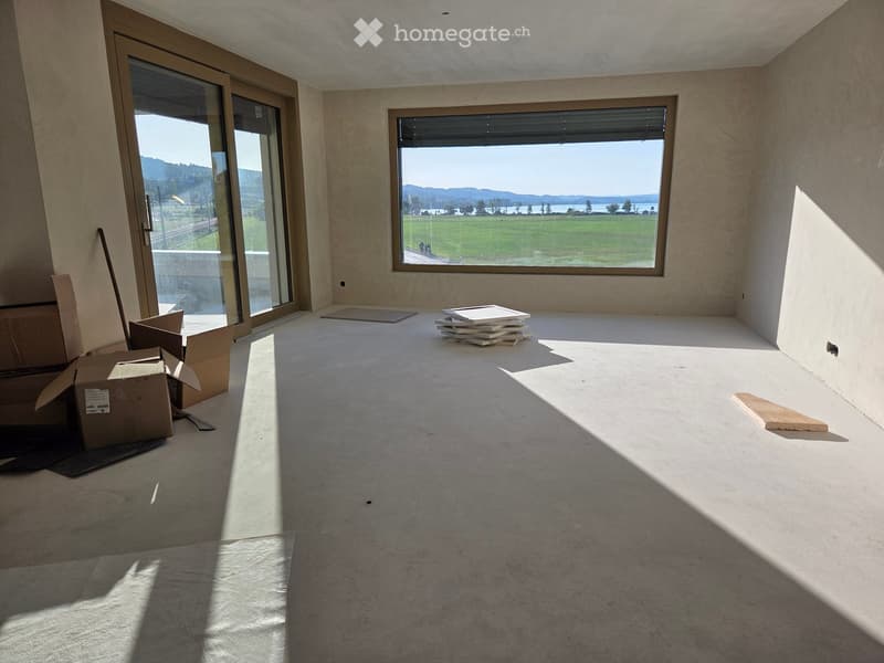 Empty room with bare walls, concrete floor, large windows, and some cardboard boxes and a stack of boards on the floor.