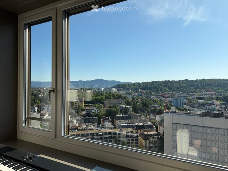 View from a window showing a cityscape with buildings, trees, and distant hills under a clear blue sky.