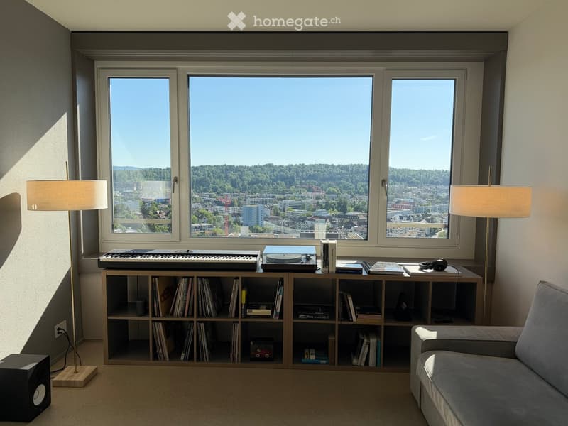 Modern living room with large windows offering a scenic city view, featuring a grey sofa, a wooden bookshelf filled with books and records, a record player, a keyboard, and two floor lamps.