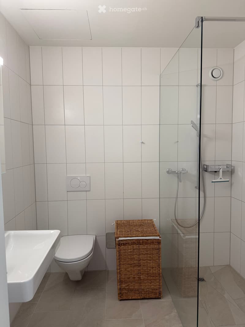 Modern bathroom with white tiled walls, a bathtub, toilet, wicker basket, and a glass-enclosed shower stall.