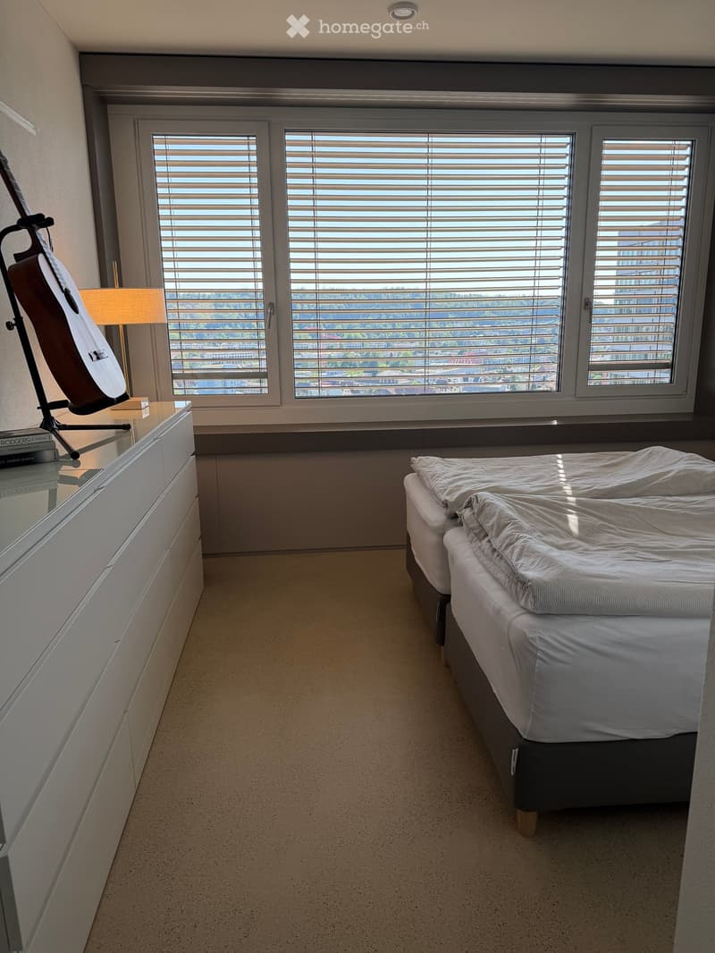 Modern bedroom with a double bed, large window with blinds offering a scenic view, a guitar on a stand, a lamp, and a mirror on a drawer unit.