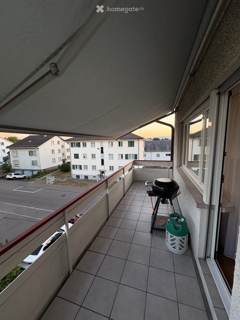 Balcony with tiled floor, metal railing, grill, and gas tank. View of parking lot and residential buildings.
