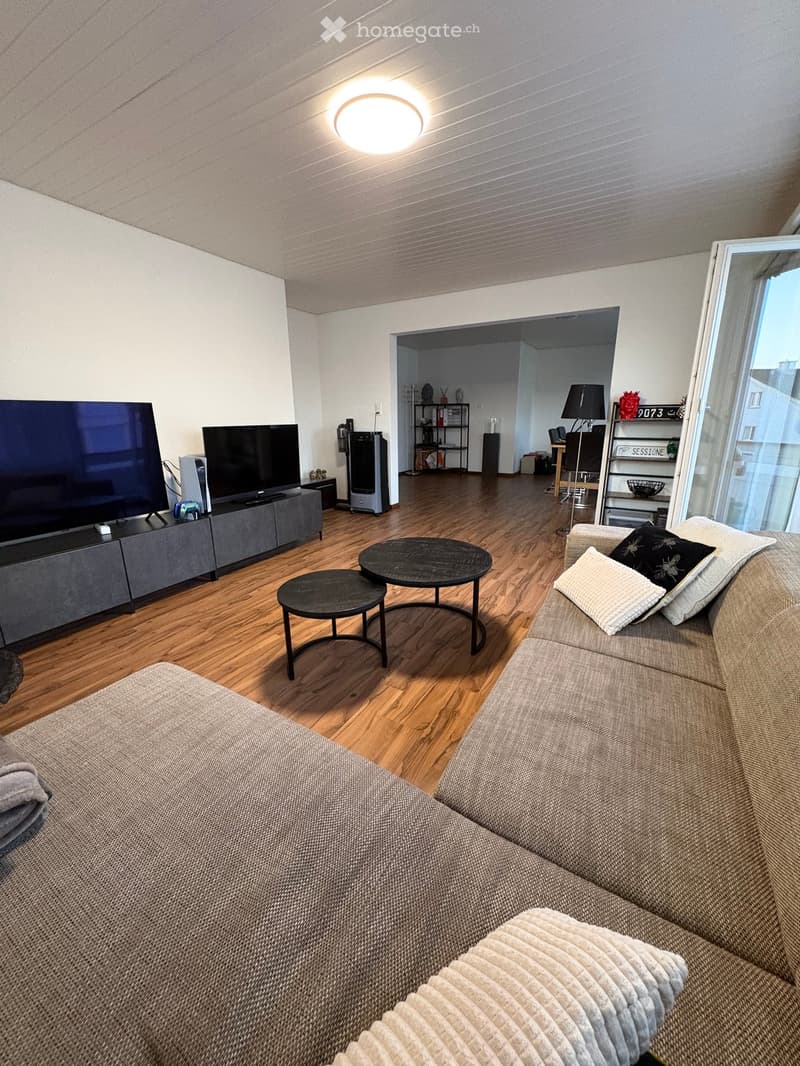 Modern living room with wooden flooring, a large sofa with pillows and a blanket, a coffee table, a stool, and a television on a stand. A lamp is visible in the background, along with a shelf and additional seating area.