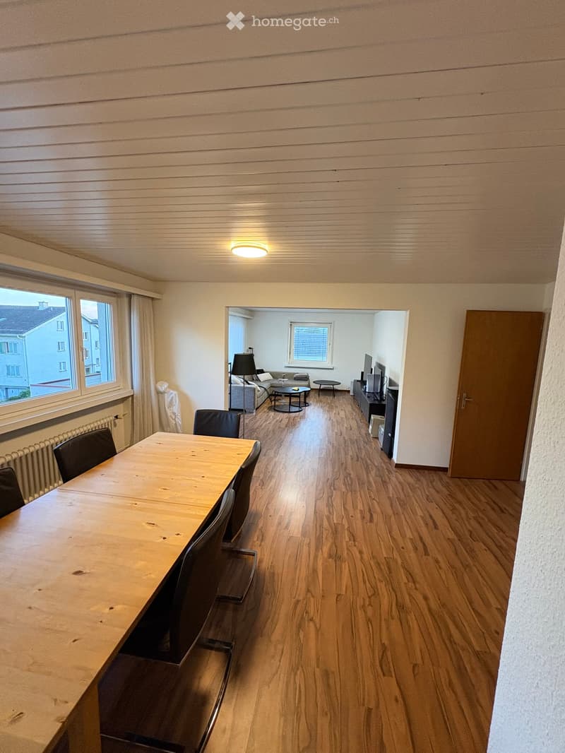 Bright, open-plan living and dining area with wooden flooring, large windows, and modern furniture including a dining table, chairs, sofa, TV, and lamp.