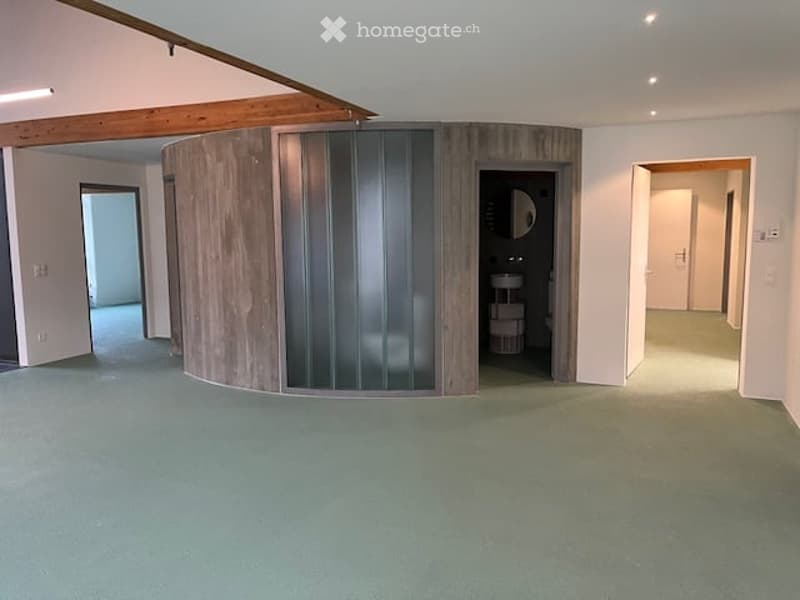 Empty room with light green carpeting, white walls, and wooden beams on the ceiling. Features a round wooden partition with glass panels and a small adjacent bathroom area.