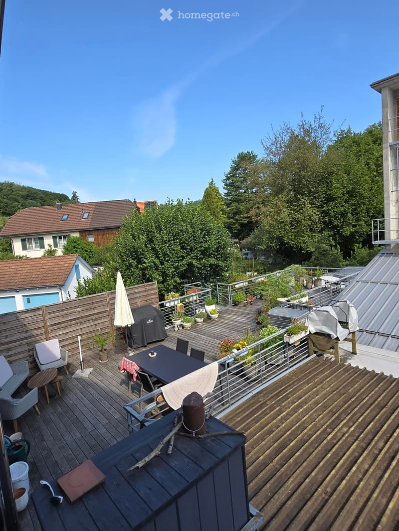 Wooden terrace with a dining table, chairs, umbrella, grill, and potted plants, surrounded by a metal railing and overlooking a green area with trees and houses.
