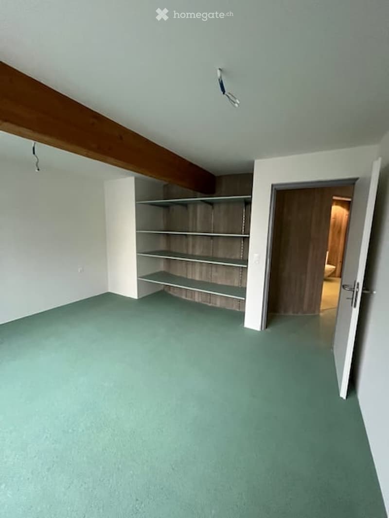Empty room with green carpet, white walls, exposed wooden beam, built-in shelving unit, and an open door leading to another room.