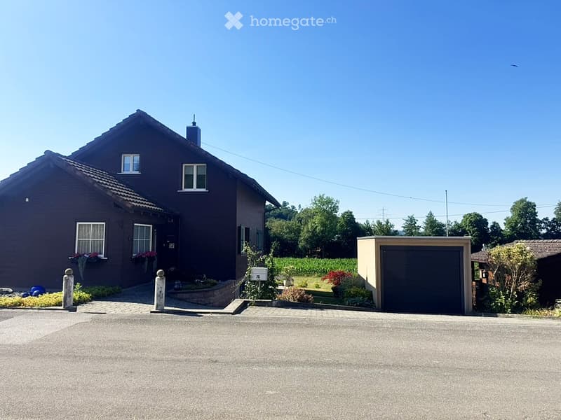 Two-story house with dark brown exterior, white-framed windows, and a garage. The house features a well-maintained garden with various plants and flowers, and a clear driveway leading to the entrance.