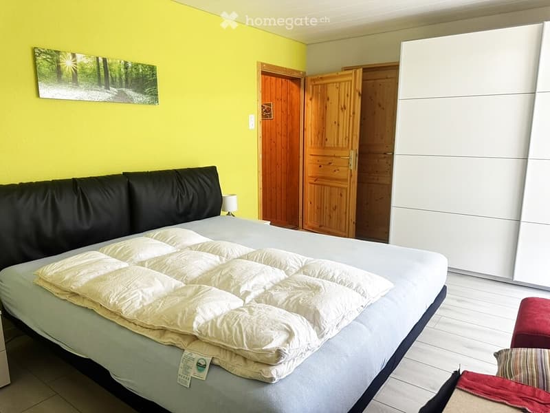 Bright bedroom with a large bed, nightstand, lamp, and a large wardrobe. The walls are painted a vibrant yellow, and a scenic forest picture hangs on the wall.