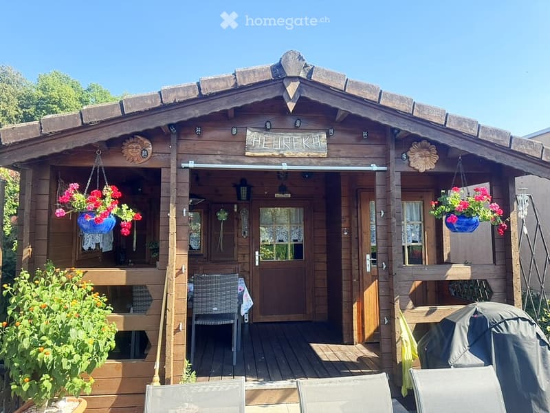 A wooden cabin with a covered porch area, featuring hanging flower pots, a table, and chairs. The entrance has a sliding door and decorative elements.
