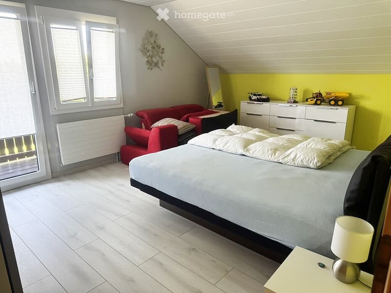 Bright bedroom with a large bed, red sofa, nightstand with lamp, and a white drawer unit with toy cars on top. The room features wooden flooring, large windows, and a slanted ceiling.