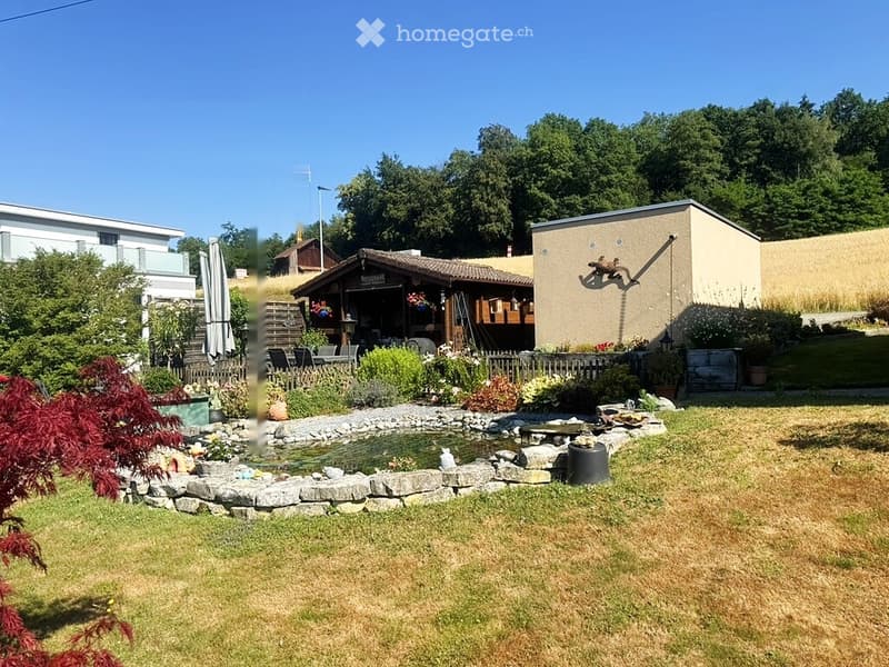 Well-maintained garden with a small pond surrounded by rocks, lush greenery, and a wooden deck area with outdoor furniture.