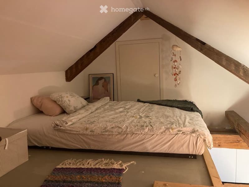 Cozy attic bedroom with slanted wooden beams, a single bed with pillows and a blanket, a picture frame on the wall, a door, and a colorful rug on the floor.