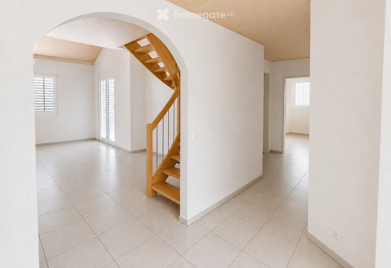 Empty hallway with white tiled flooring, white walls, and a wooden staircase leading upwards. Features include a large arched opening, a window with shutters, and a door leading to another room.