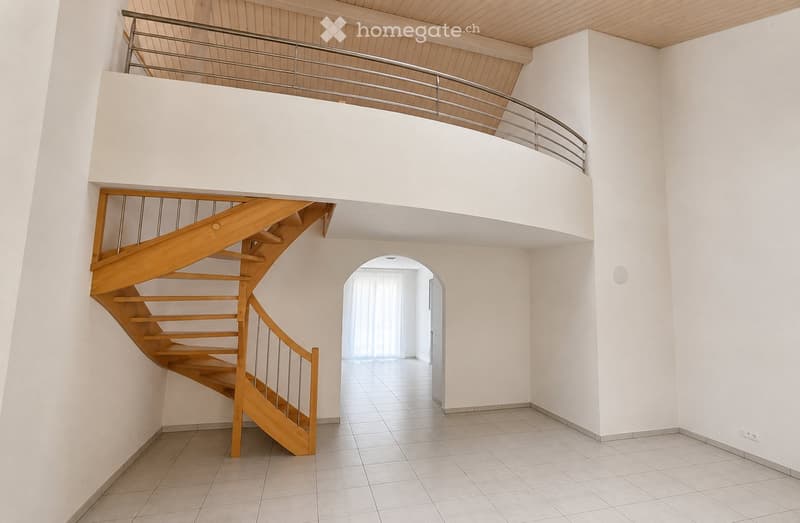 Spacious hallway with a wooden staircase leading to an upper level, white walls, tiled floor, and a balcony railing visible above.