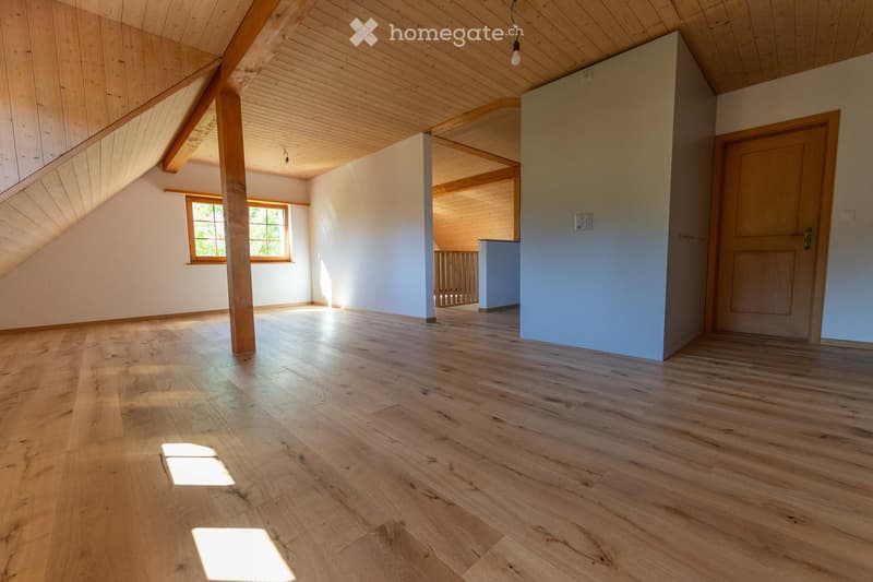 Empty room with wooden flooring, white walls, exposed wooden beams, and a window letting in natural light.
