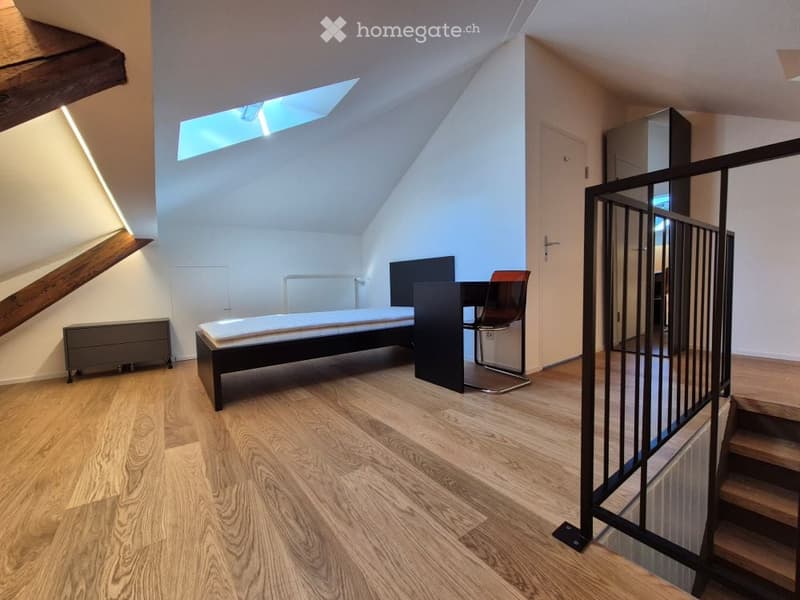 Modern attic bedroom with wooden flooring, slanted ceiling, skylight, bed, chair, table, and wardrobe.