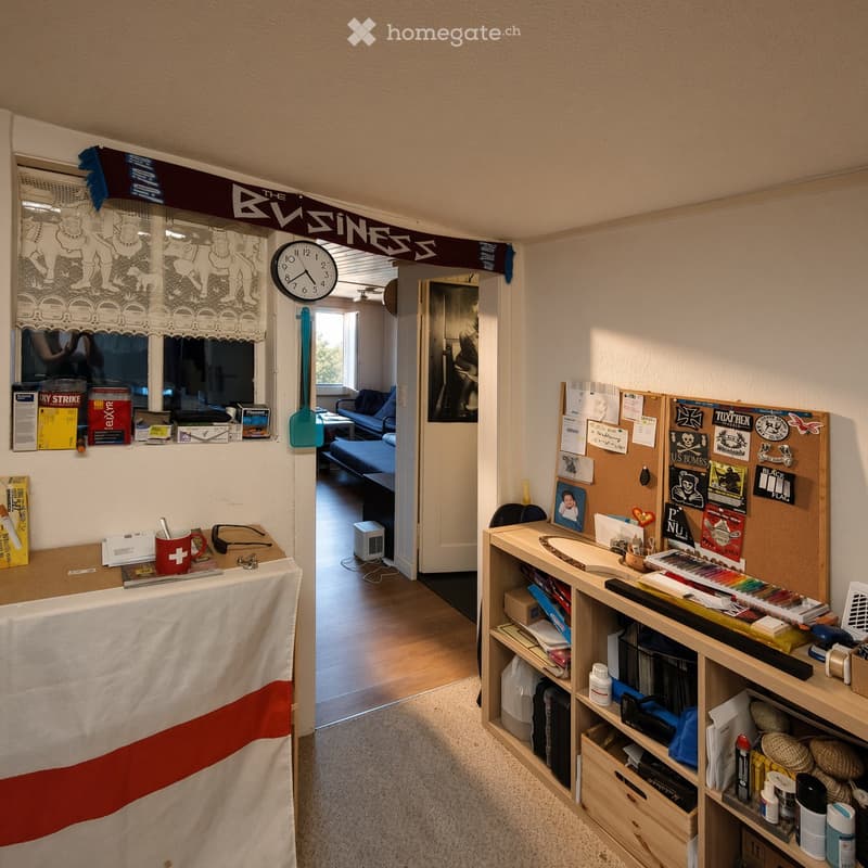 A furnished room with a table holding a cup and clock, a sofa visible through an open doorway, shelves filled with books and supplies, and a bulletin board with various items.