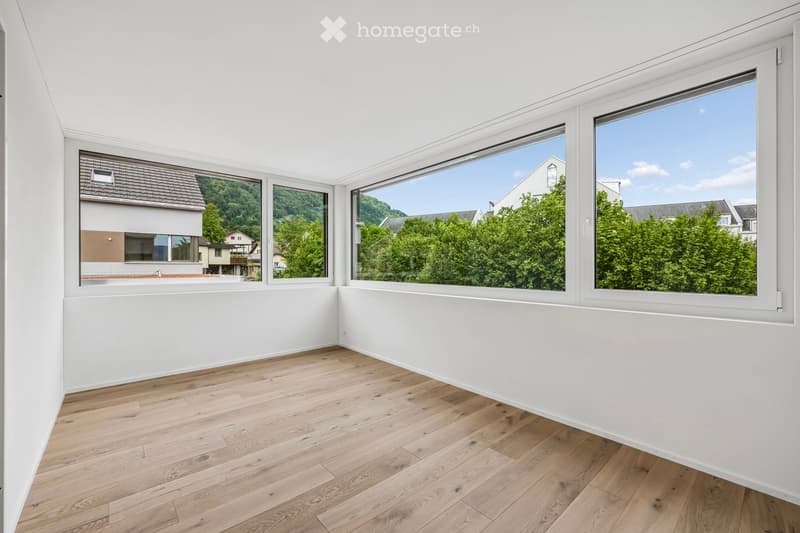 Empty room with light wooden flooring and white walls, featuring large windows offering views of greenery and neighboring buildings.