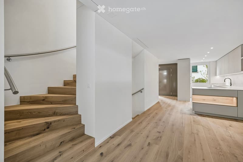 Modern hallway with wooden flooring, white walls, and a staircase leading upwards. Features a built-in kitchen area with cabinets and a sink visible in the background.