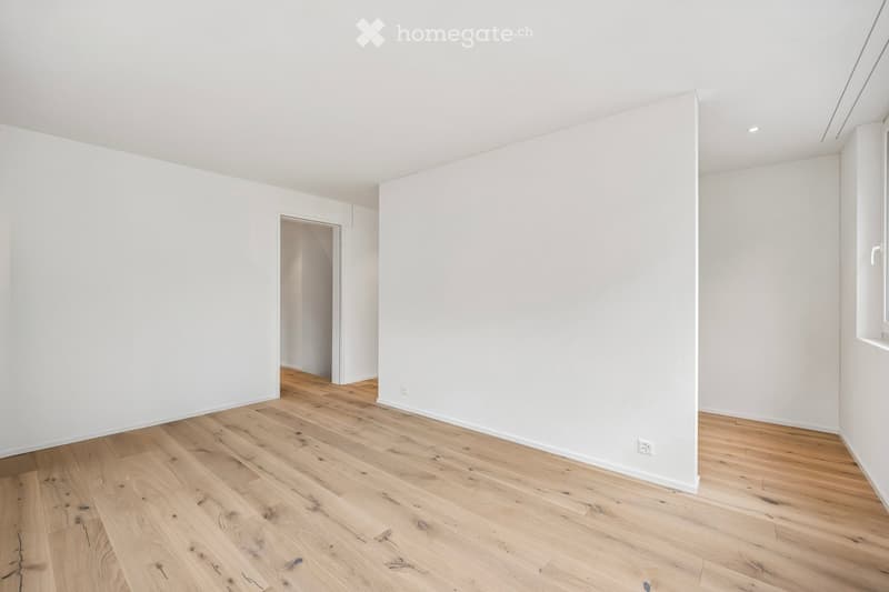 Empty room with white walls and light wooden flooring, featuring a doorway leading to another space.