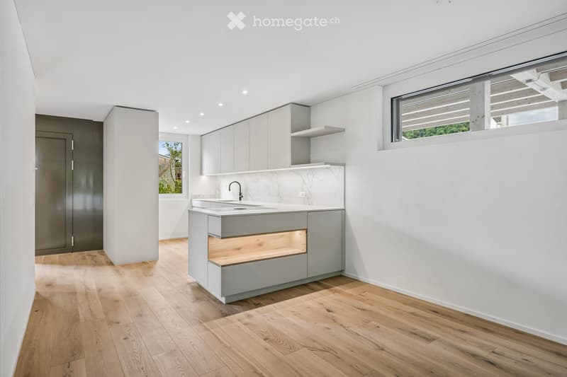 Modern kitchen with light grey cabinets, white marble countertop, and a built-in sink with a sleek faucet. The space features wooden flooring and ample natural light from large windows.