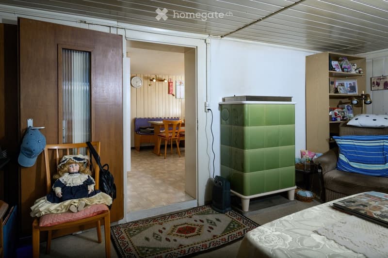A cozy living room with a sofa, a small table, a wooden chair with a doll, a green cabinet, a shelf with various items, a clock on the wall, and a heater. The room features a patterned rug and a doorway leading to another room with a dining area.