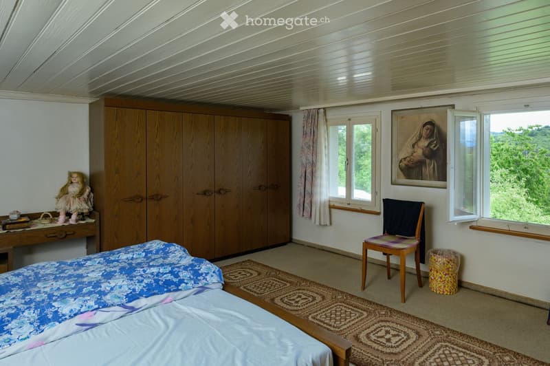 Cozy bedroom with a wooden bed, colorful bedding, a wooden wardrobe, a small table with a doll, a chair, and a large window with a scenic view.