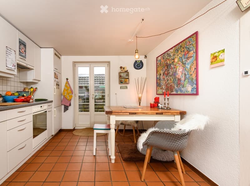 Bright kitchen and dining area with white cabinets, a wooden dining table, and colorful decor including a large abstract painting and a fruit bowl on the counter.