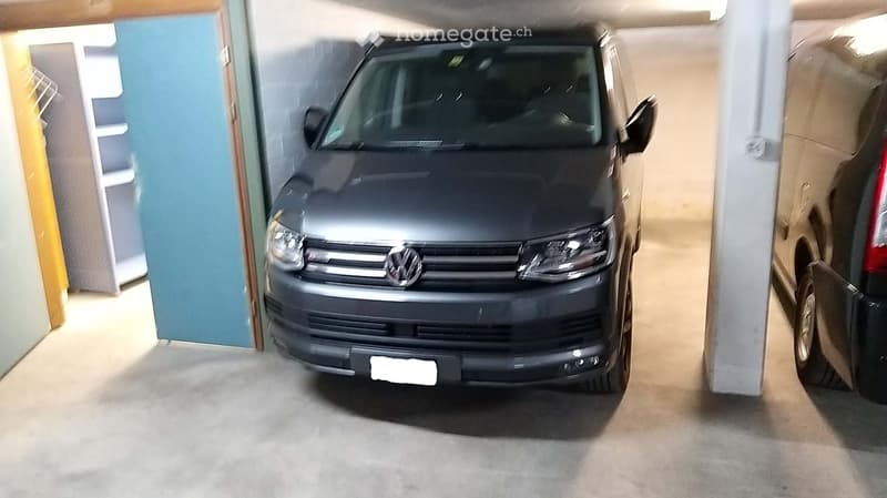 A grey Volkswagen van parked in a garage, next to a white pillar and a storage shelf, along with another car on the right